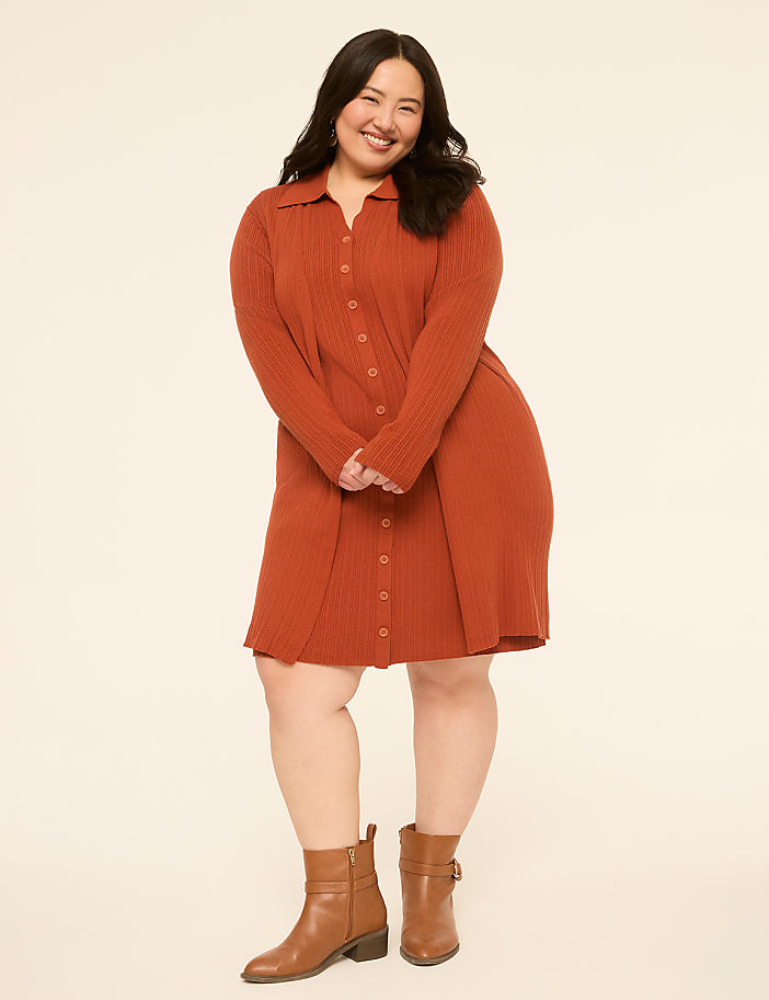A smiling woman in a rust-colored ribbed dress and brown boots stands against a plain, light-colored background.