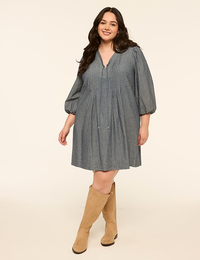 A smiling woman in a blue dress and tan boots stands against a plain, light-colored background.