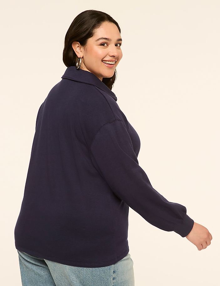 A smiling woman in a navy blue sweater and jeans is captured from the side against a plain, light-colored background.