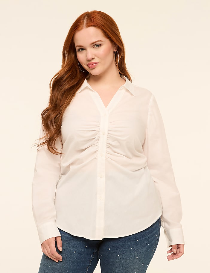 A woman with long, wavy red hair stands in the foreground wearing a white ruched blouse and jeweled jeans, against a plain, light-colored background.