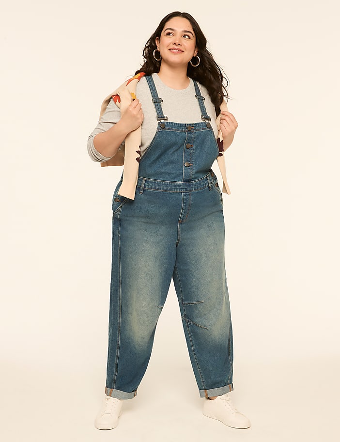 A woman in denim overalls and a sweater draped over her shoulders stands against a plain, light-colored background.
