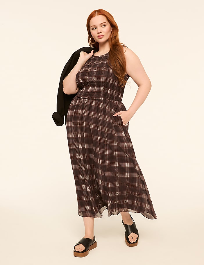 A woman in a plaid dress and sandals stands against a plain, light-colored background with a jacket draped over her shoulder.