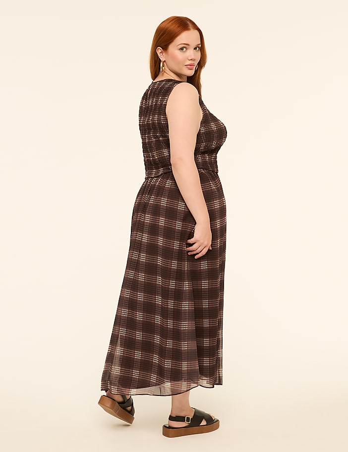 A woman with red hair in a plaid dress and platform sandals stands against a plain, light-colored background.