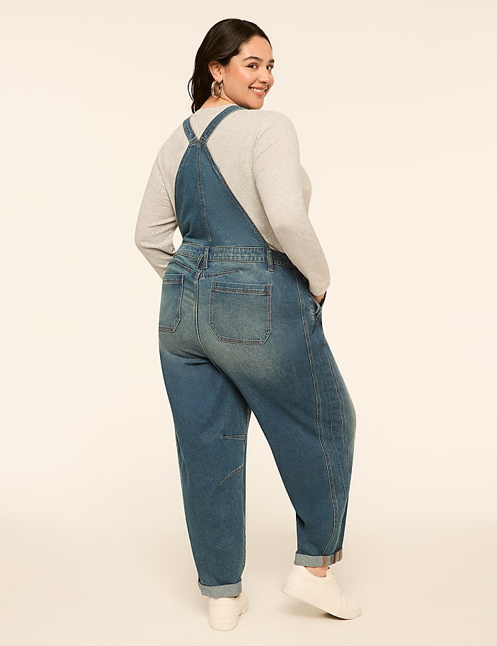A woman in denim overalls stands against a plain, light-colored background.