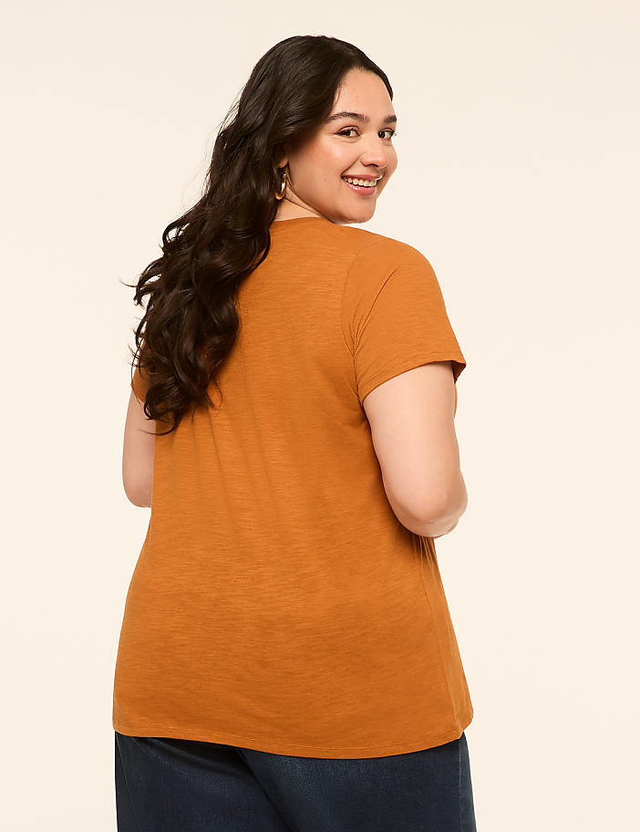 A woman in an orange t-shirt and blue jeans smiles over her shoulder against a plain cream background.