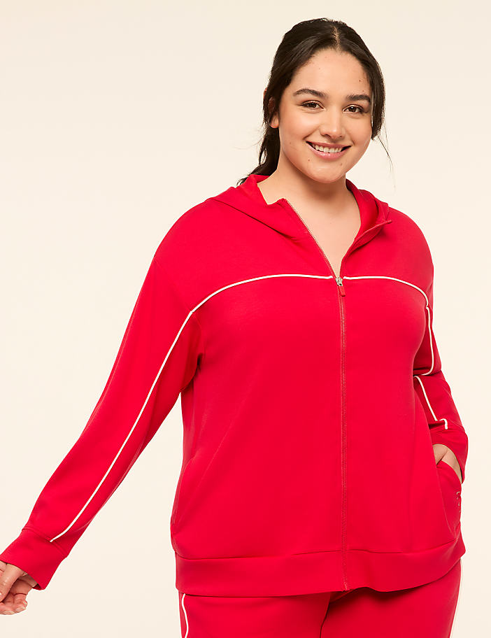 A smiling woman in a red zip-up hoodie stands against a plain, light-colored background.