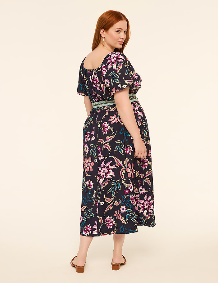 A woman with red hair wearing a floral dress stands against a plain, light-colored background.