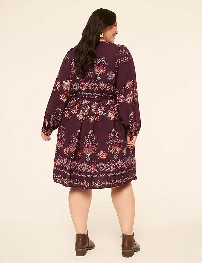 A woman in a patterned burgundy dress and brown boots stands against a plain cream background.