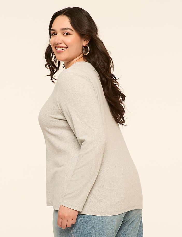 A smiling woman in a light gray long-sleeved shirt and blue jeans stands against a plain, light-colored background.