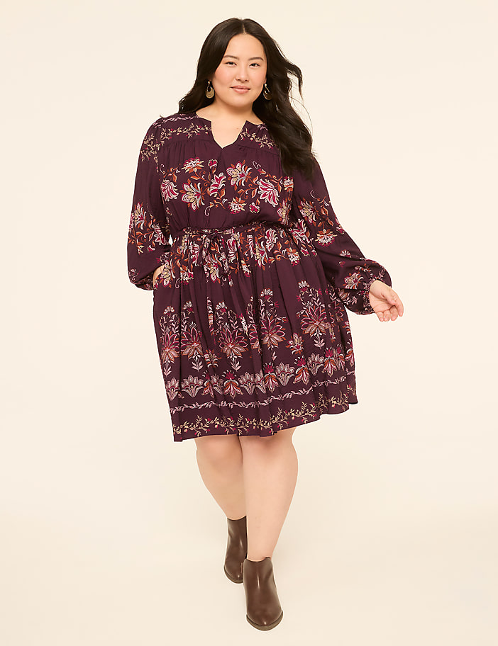 A woman in a floral print dress and brown boots stands against a plain, light-colored background.
