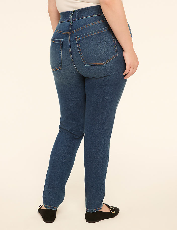 A person wearing blue jeans and black flats stands against a plain cream background.