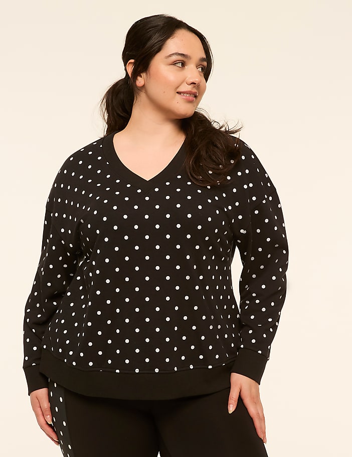 A woman with dark hair pulled back in a ponytail, wearing a black and white polka dot v-neck sweatshirt, stands against a plain, light-colored background.