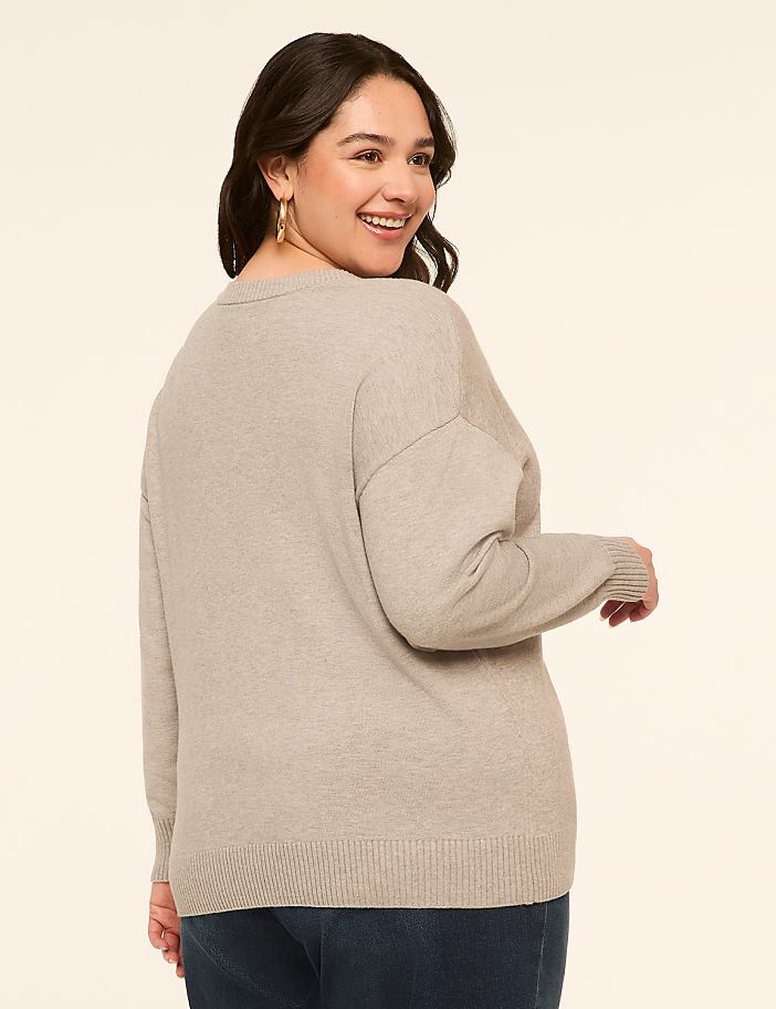 A woman in a light gray sweater and dark jeans smiles over her shoulder against a plain, light-colored background.