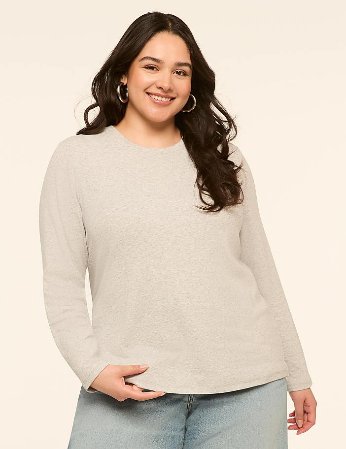 A smiling woman with dark hair and hoop earrings in a light gray long-sleeved shirt and blue jeans stands against a plain, light beige background.