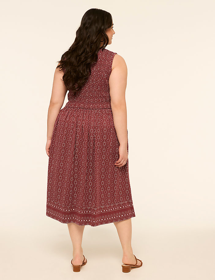 A woman in a patterned dress stands against a plain, light background.