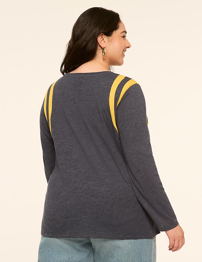 A woman in a dark blue long-sleeved shirt with yellow stripes on the shoulders stands against a plain, light-colored background.