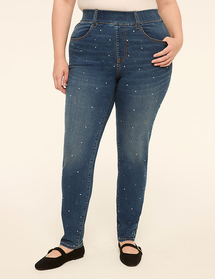 A person wearing studded denim jeans and black ballet flats stands against a plain, light-colored background.