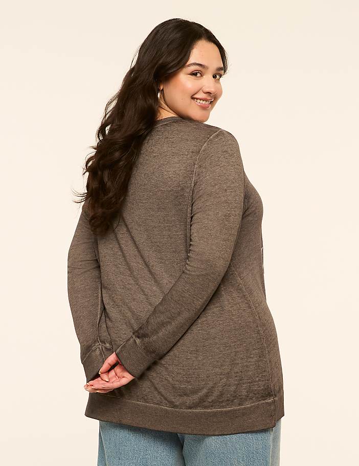 A woman with dark hair and a brown sweater stands with her hands behind her back against a plain, light-colored background.