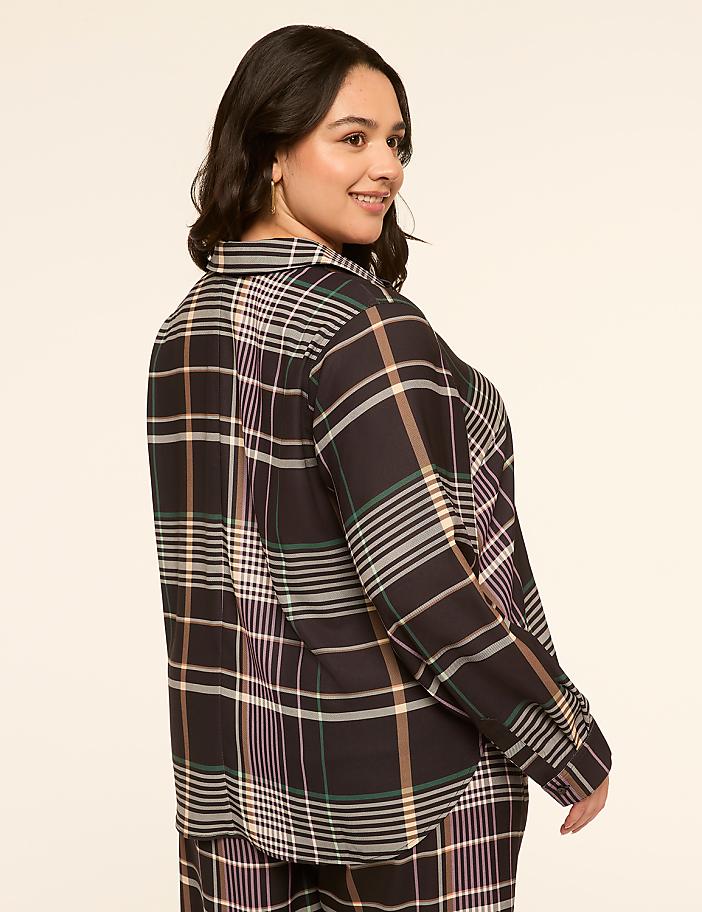 A woman in a plaid shirt and pants smiles over her shoulder against a plain, light-colored background.