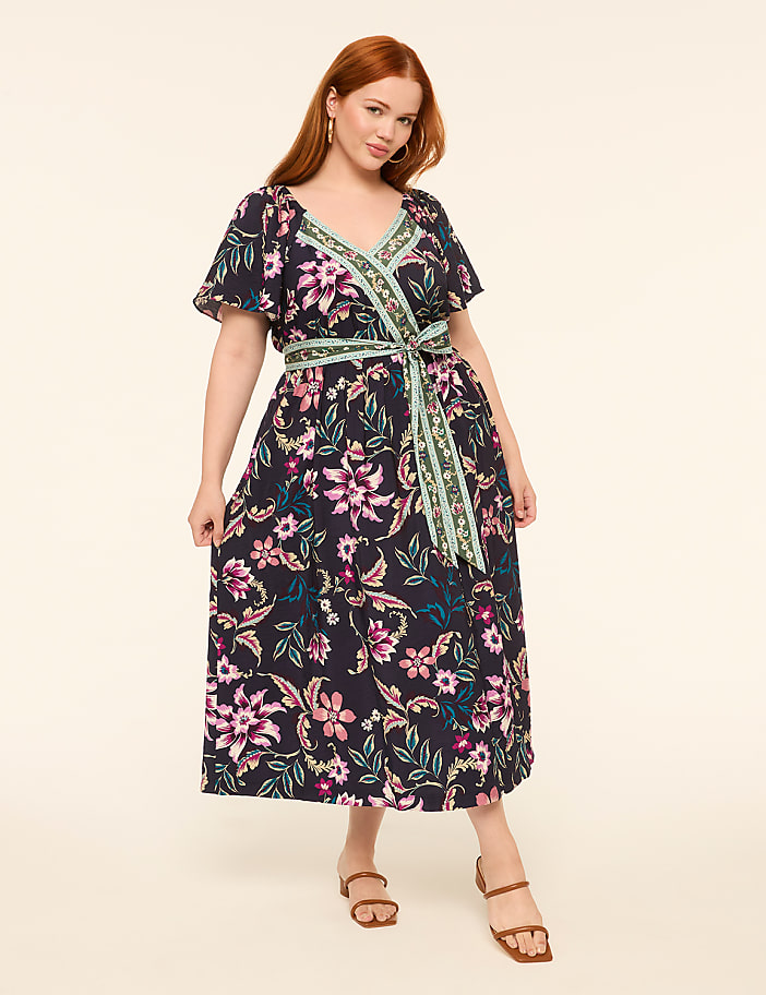 A woman in a floral wrap dress stands against a plain, light-colored background.