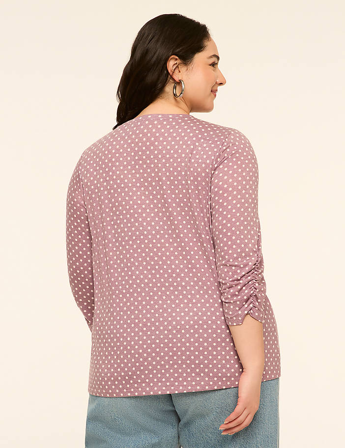 A woman in a polka dot shirt and jeans stands against a plain, light-colored background.