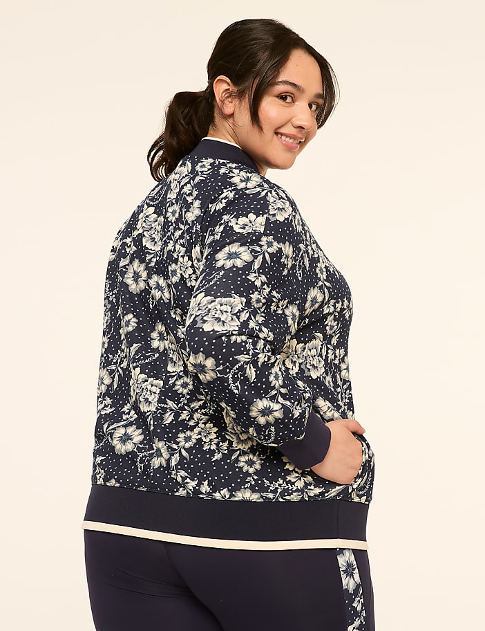 A woman in a floral bomber jacket and navy pants smiles over her shoulder against a plain, light background.