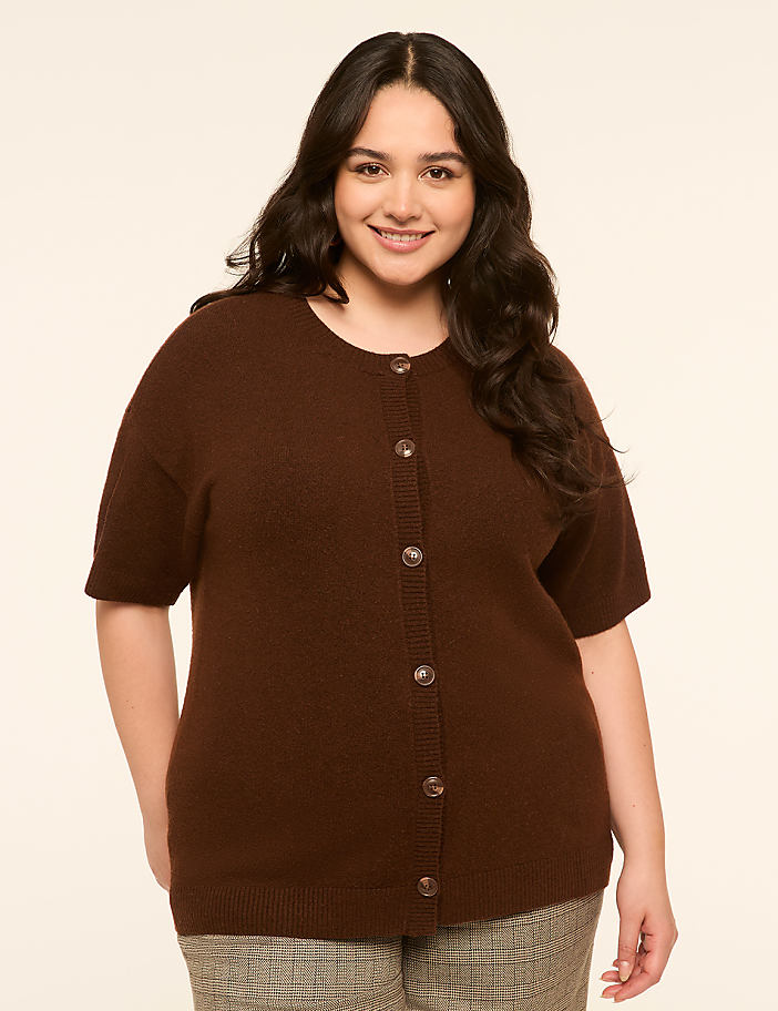 A smiling woman in a brown cardigan stands against a plain, light-colored background.
