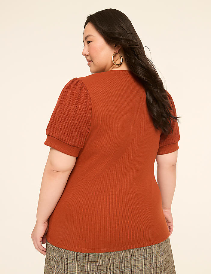 A woman in a rust-colored puff-sleeve top and plaid skirt stands against a plain, light-colored background.