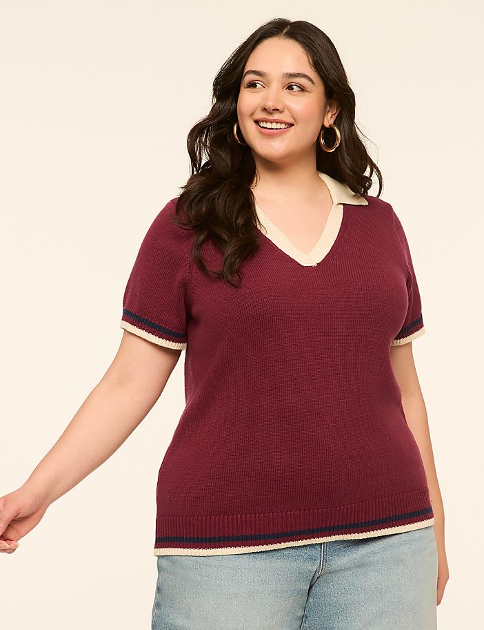 A smiling woman in a maroon collared shirt and denim shorts stands against a plain, light-colored background.