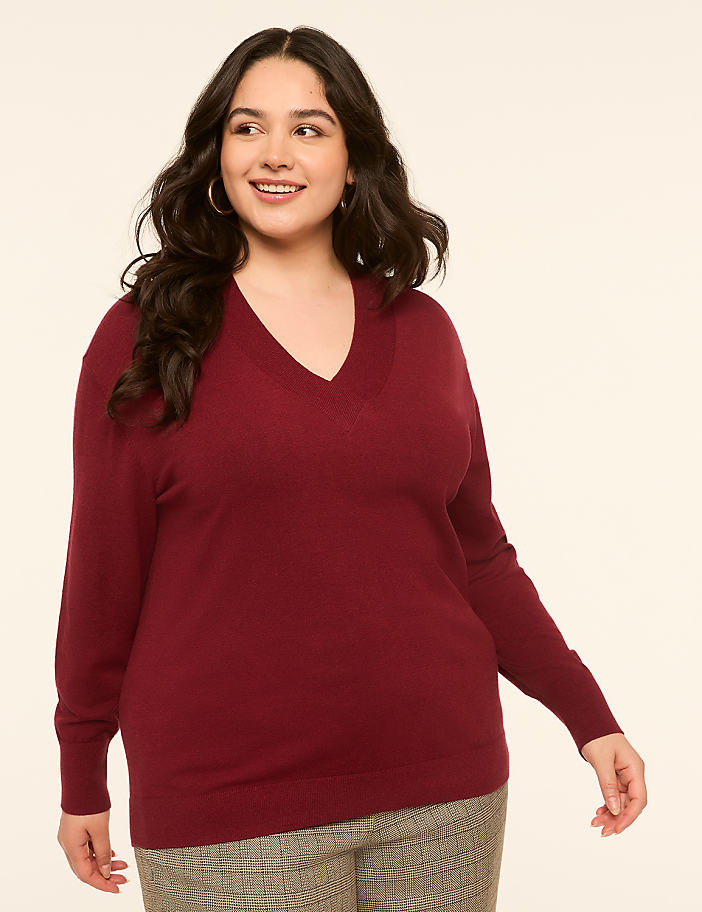 A smiling woman in a maroon v-neck sweater and plaid pants stands against a plain, light-colored background.