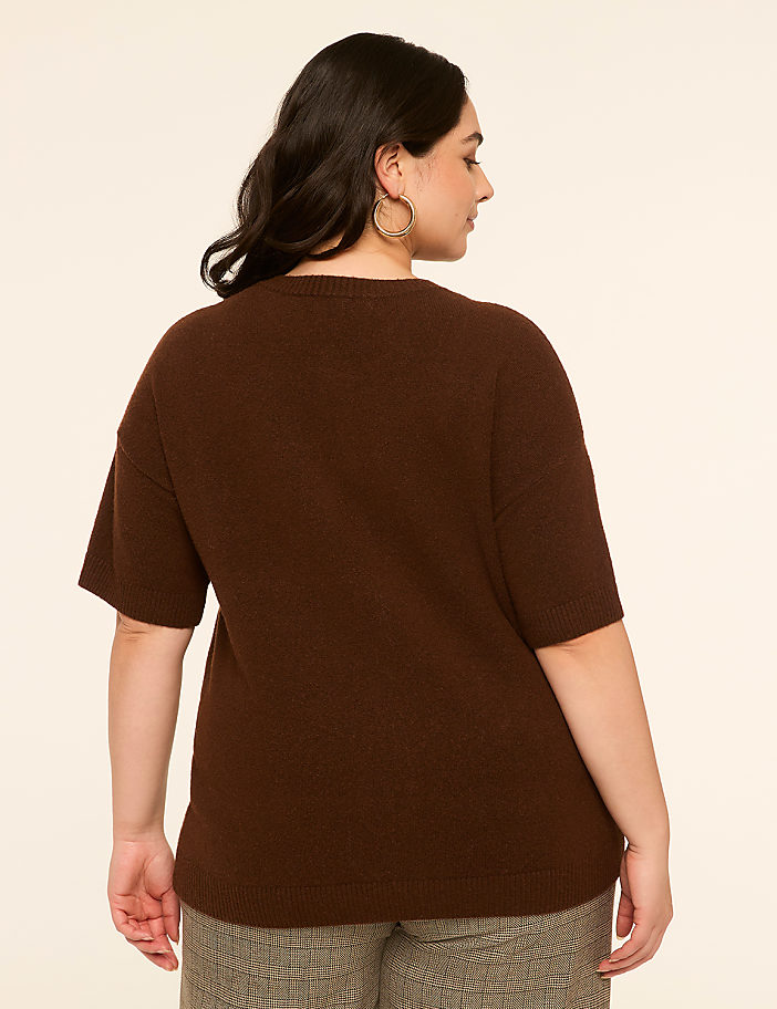 A woman in a brown sweater and patterned pants stands against a plain, light-colored background.