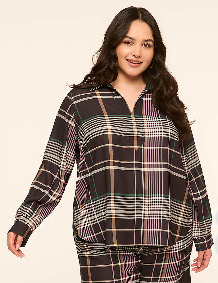 A smiling woman with dark, wavy hair poses in a dark, plaid shirt and matching pants against a plain, light-colored background.