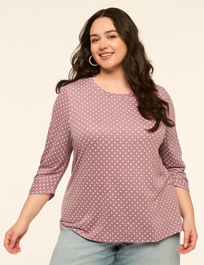 A smiling woman with dark, wavy hair and hoop earrings, wearing a purple polka-dot shirt and light blue jeans, stands against a plain, light-colored background.
