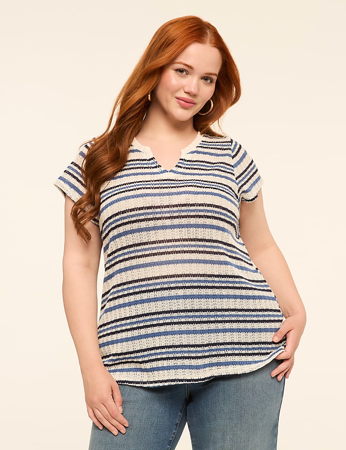 A woman with red hair wearing a striped shirt and jeans stands against a plain, light-colored background.