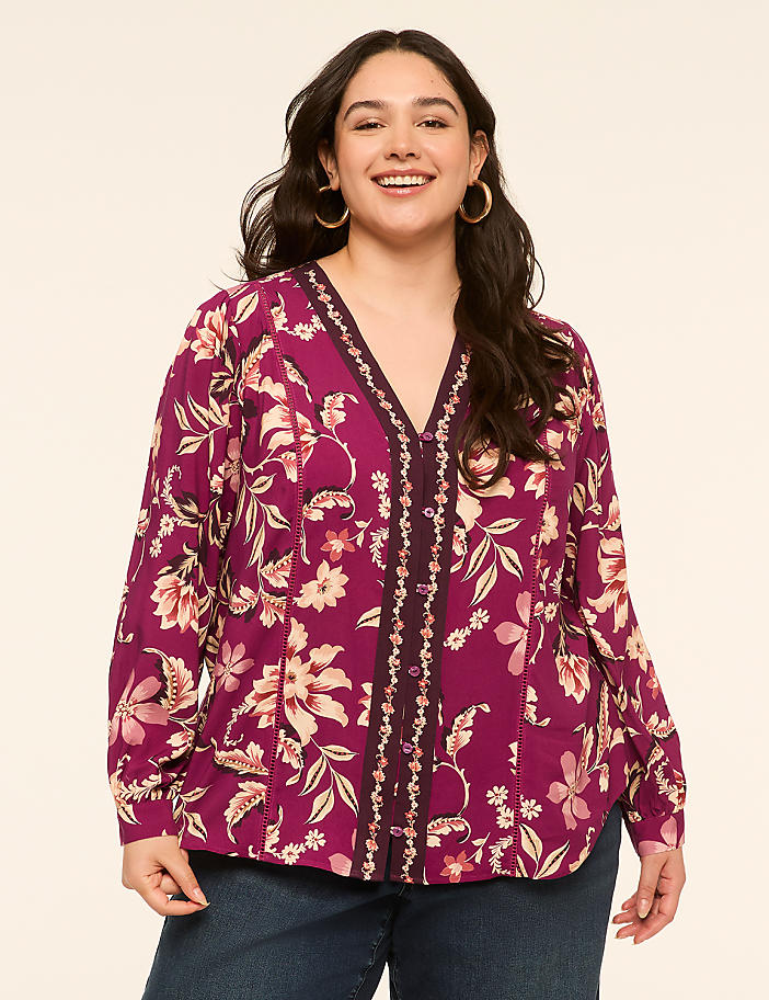A smiling woman with dark hair and hoop earrings poses in a floral print blouse and jeans against a plain, light-colored background.