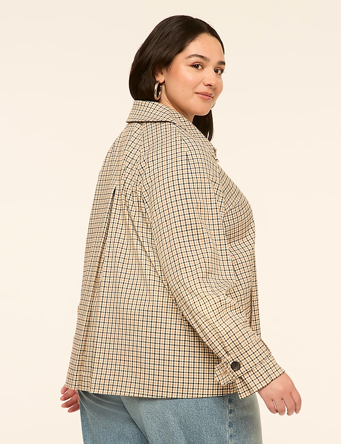 A woman in a checkered jacket and jeans stands against a plain, light-colored background.
