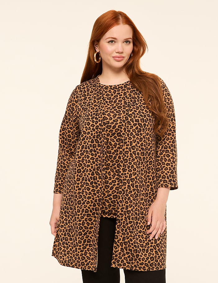 A woman with long red hair and large hoop earrings stands in the foreground wearing a leopard print jacket over black pants, against a plain, light-colored background.