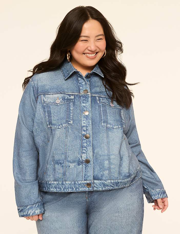 A smiling woman in a denim jacket and jeans stands against a plain, light-colored background.