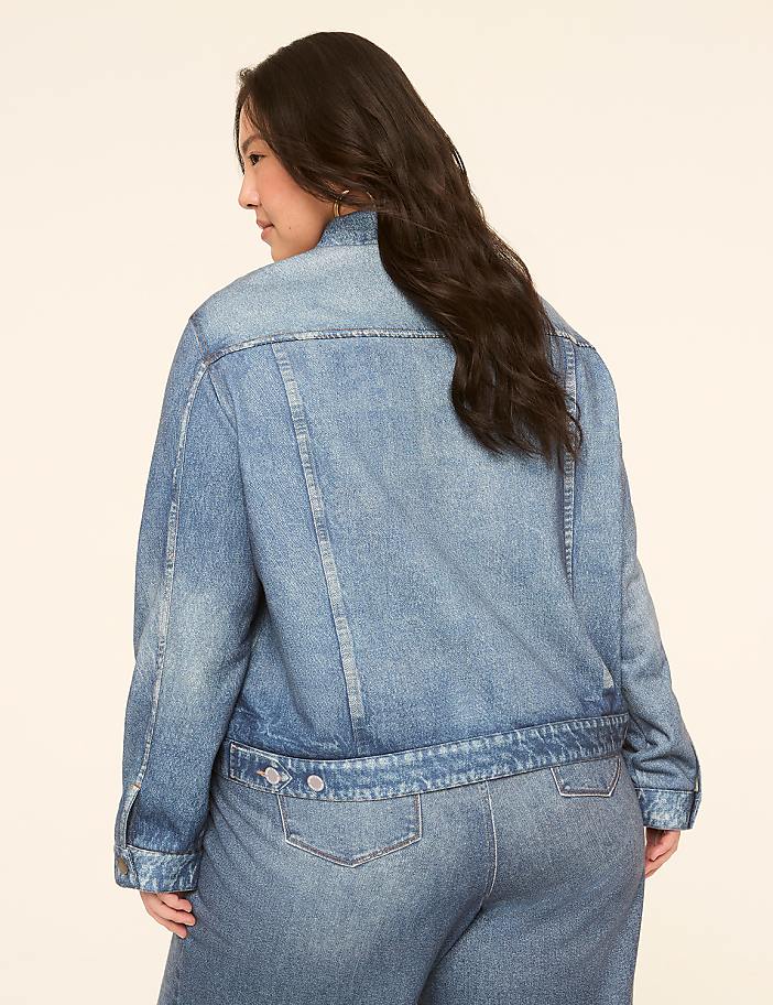 A woman in a denim jacket and jeans stands against a plain, light-colored background.