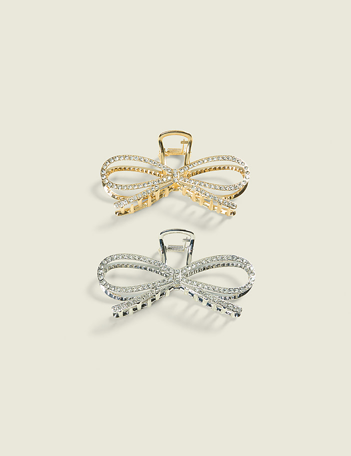 Two sparkling bow-shaped hair clips, one gold and one silver, rest on a plain, light-colored background.