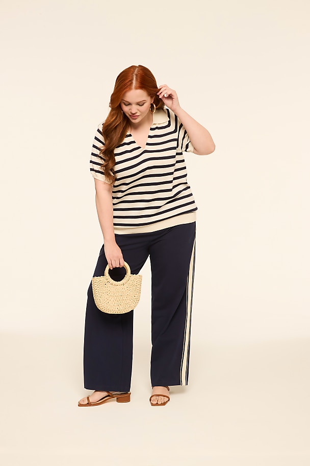 A woman with red hair stands in the foreground holding a straw bag, while the background is a plain, light-colored studio backdrop.