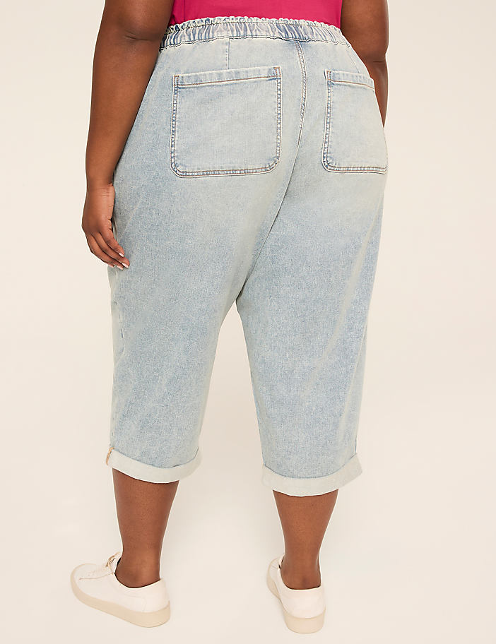 A person wearing light-wash denim capri pants with white sneakers stands against a plain, light-colored background.