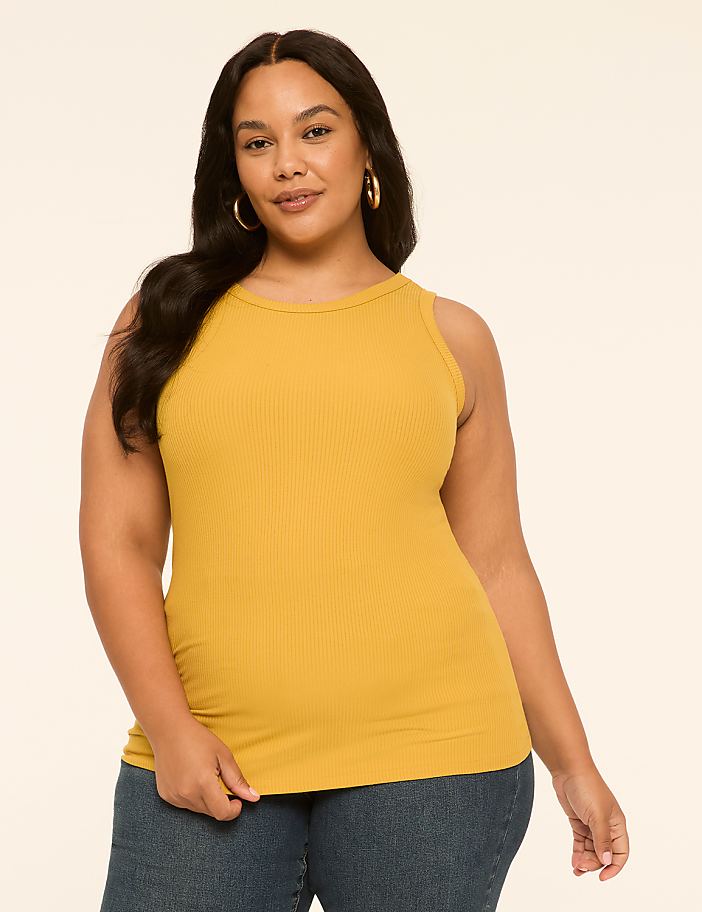 A woman in a mustard yellow ribbed tank top and blue jeans stands against a plain, light-colored background.