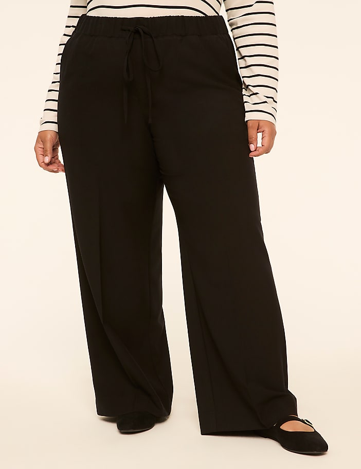 A person in black wide-leg pants and a striped shirt stands against a plain, light-colored background.