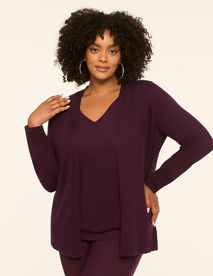 A woman with curly dark hair poses in a plum-colored cardigan and matching top against a plain cream background.