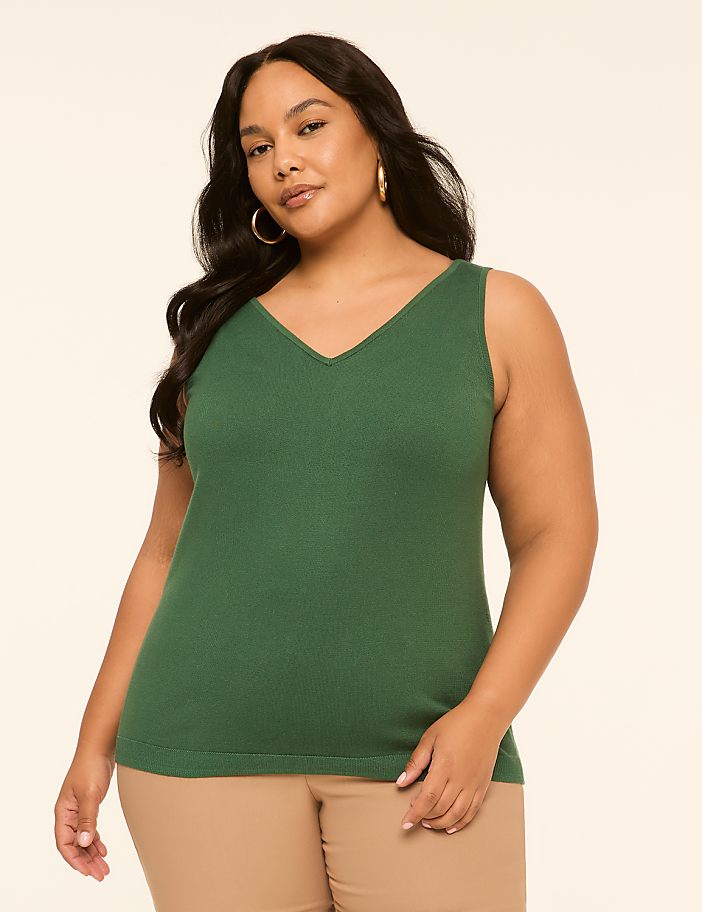 A woman in a green v-neck tank top and tan pants poses against a plain cream background.
