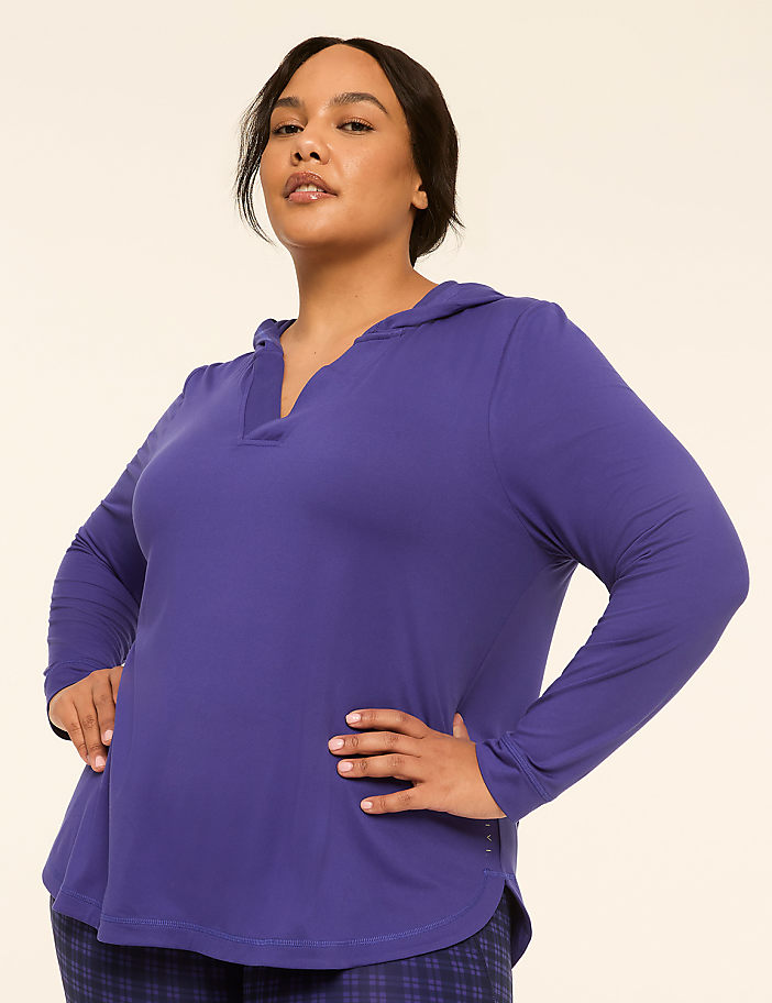 A woman in a purple hooded shirt and plaid pants stands with her hands on her hips against a plain, light-colored background.