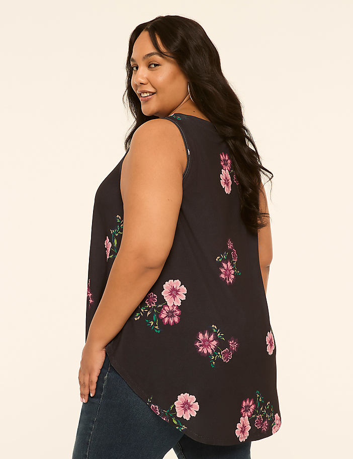 A woman in a floral tank top and jeans stands against a plain, light-colored background.