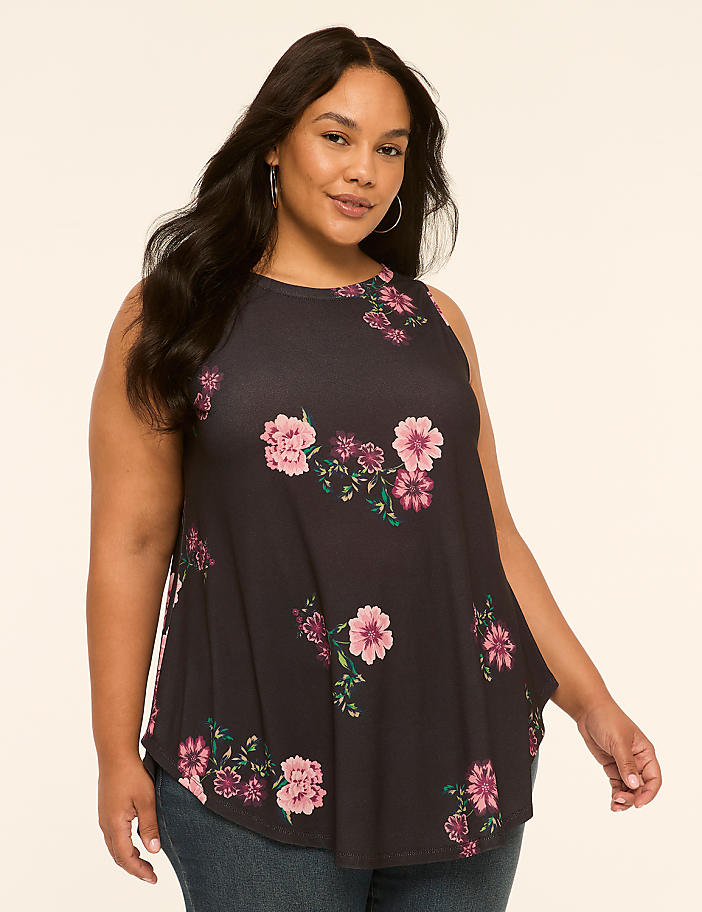 A woman with dark hair and a floral tank top stands against a plain, light-colored background.