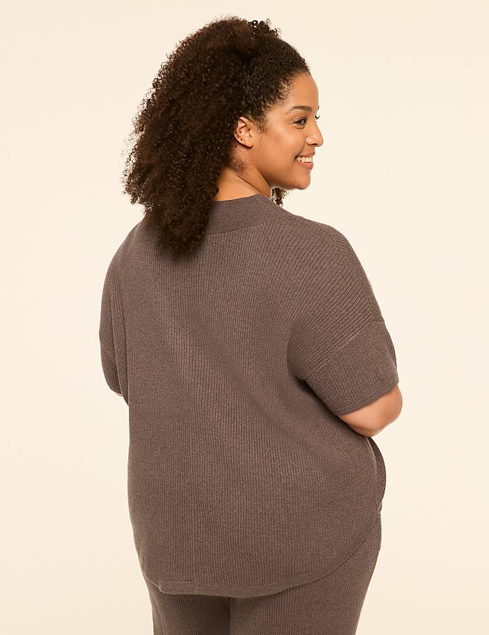 A woman with curly dark hair, wearing a gray ribbed sweater and pants, smiles over her shoulder against a plain cream background.